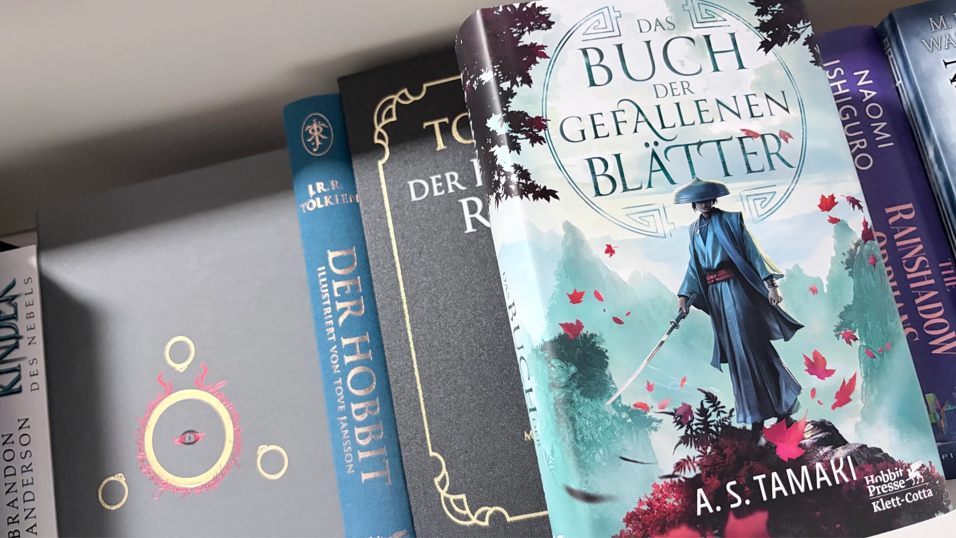 Foto von einem Bücherregal. Hevorgehoben ist das Buch "Das Buch der fallenden Blätter" auf dem Cover ist oben der Titel zu lesen und mittig steht eine Frau mit einem Katana auf einer kleinen Erhöhung.